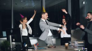 Happy young people office workers in suits are dancing at company party wearing party hats and blowing horns. Birthday cake and gift box are visible on desk.